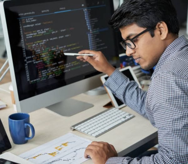 Serious Indian programmer in eyeglasses pointing at computer monitor and checking the statistics of website in document while working at office